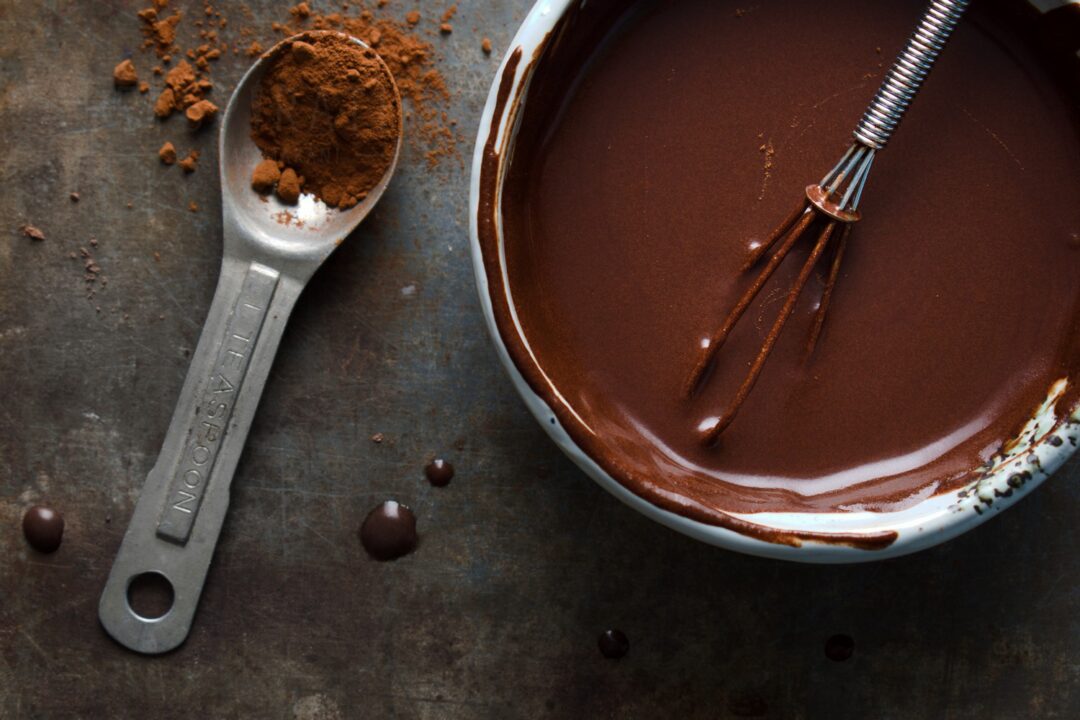 Een schaaltje met chocoladesaus met een sausklopper, ernaast een theelepel cacaopoeder