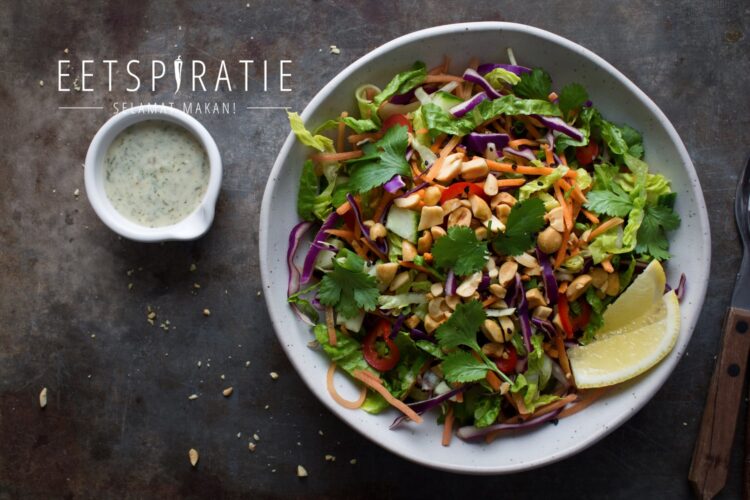 Een bord salade met wortel, rode kool, spitskool, komkommer, Romaine sla, buisuitjes, pinda's en verse koriander. Geserveerd met partjes citroen en een frisse dressing in een kannetje